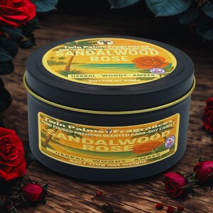 A black tin candle with a gold rim and label featuring the text "Sandalwood Rose". The label includes a graphic of a rose and palm trees. The candle is 6 oz (170g) and is surrounded by red roses.