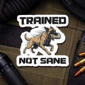 May include: A sticker featuring a tan and black dog carrying a training object in its mouth. The text "TRAINED" is above the dog, and "NOT SANE" is below. The sticker has a white border.