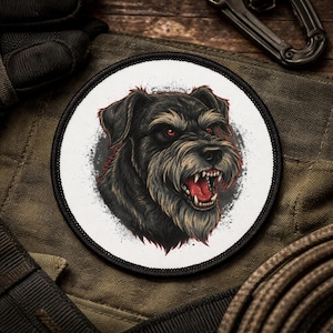 May include: Round patch featuring a detailed illustration of an angry dog with red eyes and an open mouth, baring its teeth. The dog is black and tan, set against a white background with a black border. The patch is on a green fabric.