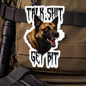 May include: A white sticker with the text "TALK SHIT GET BIT" and a snarling German Shepherd dog illustration. The dog is brown and black with red eyes and an open mouth. The sticker is attached to a green tactical bag.