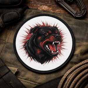 May include: A round patch featuring a snarling black dog with red accents and glowing eyes. The dog's open mouth reveals sharp white teeth, set against a white background with a red burst design. The patch has a black border.