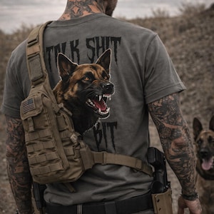 May include: Grey t-shirt with the text "TALK SHIT BIT" and a snarling German Shepherd dog graphic. A tactical sling bag with an American flag patch is worn across the chest. Another dog is visible in the background.