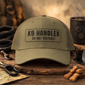 May include: An olive green trucker hat with the text "K9 Handler Do Not Distract" in a black rectangular box. The hat has a mesh back and a curved brim. Accessories include dog treats, a leash, and sunglasses.
