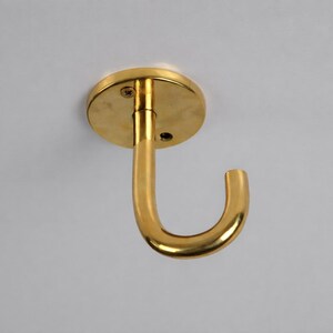 Puede incluir: Un gancho de techo dorado con una base circular y un gancho curvo. El gancho es de metal y tiene un acabado brillante. La base tiene dos orificios para tornillos para el montaje.