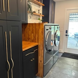 May include: A modern laundry room with black cabinets and gold hardware. A stainless steel washer and dryer are set into a wooden cabinet. The room has a wooden countertop and open shelving with decorative items.