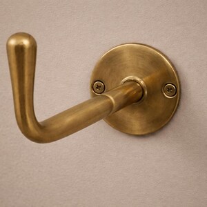 May include: A brass wall hook with a curved arm and a rounded end. The hook is attached to a circular base with two screws. The finish is a warm, antique gold color.