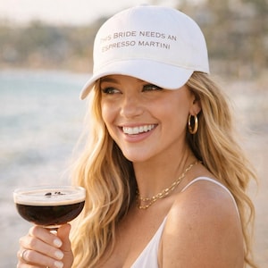 May include: A white baseball cap with the text "THIS BRIDE NEEDS AN ESPRESSO MARTINI" in black lettering. The cap is worn by a person holding a cocktail. The cocktail is in a clear glass with a dark liquid and coffee beans.