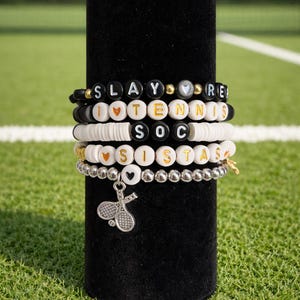 May include: A collection of beaded bracelets with various phrases and charms. The bracelets are black, white, and gold, with the words "SLAY," "RE," "I LOVE TENNIS," "SOC," and "SISTAS" spelled out. A tennis racket charm hangs from one bracelet. The bracelets are displayed on a black cylinder.