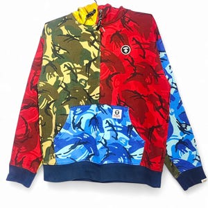 May include: A color-blocked hoodie with a camouflage pattern in red, green, yellow, and blue. The hoodie has a kangaroo pocket and a navy blue hem and cuffs. A small logo is visible on the chest and pocket.
