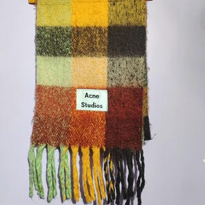 May include: A colorful scarf with a checkered pattern in shades of yellow, orange, brown, and green. The scarf has a fringed edge with green, yellow, and black tassels. The label reads "Acne Studios".