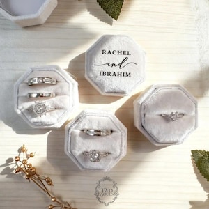 Peut inclure: Plusieurs boîtes à bagues en velours gris, chacune contenant une bague en argent. L'une des boîtes est personnalisée avec les noms "Rachel et Ibrahim". Les boîtes sont disposées sur une surface en bois clair, avec une brindille de fleurs décoratives dorées.