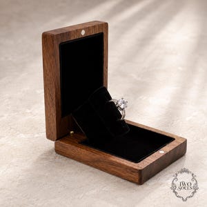 May include: A dark brown wooden ring box with a hinged lid and a black velvet interior insert. A silver-toned ring with a prominent round clear stone is positioned on the velvet display. The text TWO SOULS is printed in the bottom right corner.