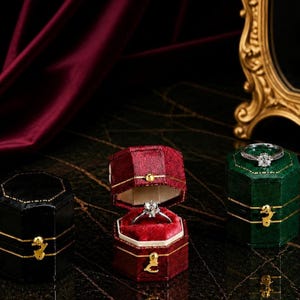 Puede incluir: Tres joyeros hexagonales en negro, rojo y verde, cada uno con detalles dorados. La caja roja está abierta, mostrando un anillo de diamantes. La caja verde también contiene un anillo de diamantes. Las cajas están sobre una superficie oscura.