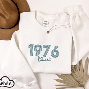 May include: White sweatshirt with the year "1976" in teal blue, with the word "Classic" underneath. A brown hat and a gold necklace are also visible. The sweatshirt is folded, with the sleeve and cuff visible.