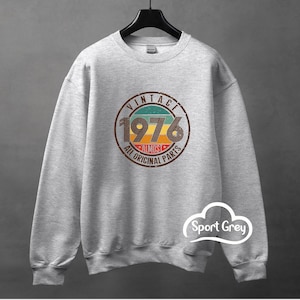 Può includere: Felpa grigio sport con un design vintage. Il design presenta una grafica circolare con la scritta "VINTAGE 1976 Almost All Original Parts" in marrone, arancione e turchese. La felpa è appesa a una gruccia nera.