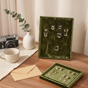 Puede incluir: Exhibición de joyas, incluyendo anillos y pendientes, presentados en bandejas de terciopelo verde. Los anillos presentan diseños de estrellas, lunas y flores. Una cámara vintage, una taza y un sobre también están sobre la superficie de madera.