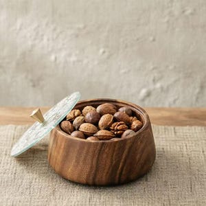 Może przedstawiać: Drewniana miska wypełniona różnymi orzechami, w tym orzechami włoskimi i pekan, ze szklaną pokrywką. Pokrywka ma złoty uchwyt. Miska stoi na neutralnej powierzchni.