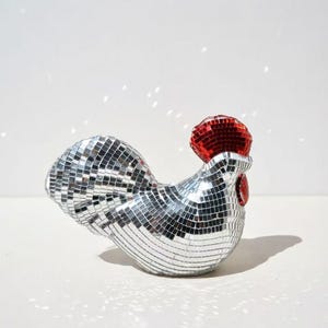 May include: A decorative rooster figurine covered in small, reflective mirrored tiles. The rooster has a red comb and wattle. The body is silver, and the comb and wattle are red. The figurine is a unique home decor item.