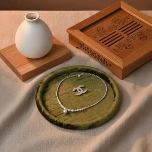 May include: A still-life composition featuring a white vase on a wooden stand, a decorative wooden box, and a green velvet tray holding a pearl necklace and a matching pendant. The jewelry has a silver finish.