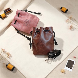 May include: A pink drawstring bag with a camera graphic and the text Oliver Smith, and a brown drawstring bag with a camera graphic and the text Emma's Camera. Both bags feature black cords, metal eyelets, and a detachable black shoulder strap.