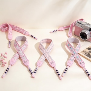 May include: Three camera straps in shades of pink, white, and light blue with woven patterns. Each strap features pink leather-like ends with the name Ruby printed in black. One strap is attached to a pink and silver camera with the name Ellie printed.