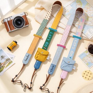 May include: Two camera straps with attached small pouches. The left strap is yellow and green with a checkered pattern and the name Amelia. The right strap is blue and pink with stripes and the name Olivia. Both feature the text LET'S GO.