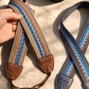 May include: Two camera straps featuring a herringbone weave pattern in beige and dark gray. Each strap has a central blue band with orange embroidery and brown or gray leather end tabs embossed with the names William and James.