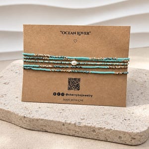May include: A set of four delicate beaded bracelets featuring turquoise and metallic gold seed beads, with a single white freshwater pearl centred on one strand. The bracelets are displayed on a rectangular brown card labelled OCEAN LOVER.