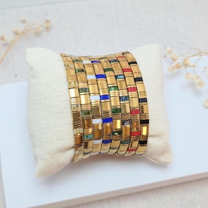 Peut inclure: Un ensemble de sept bracelets empilés composés de perles rectangulaires en métal doré alternant avec des segments colorés en blanc, bleu, vert, turquoise, rouge et noir, disposés en rangée verticale sur un petit coussin de présentation en tissu écru.