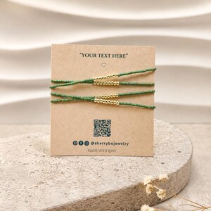 Peut inclure: Deux bracelets fins composés de petites perles vertes avec une section centrale de perles métalliques dorées, présentés sur une carte carrée beige. La carte porte le texte "YOUR TEXT HERE", un QR code, des icônes de réseaux sociaux et "MADE WITH LOVE".
