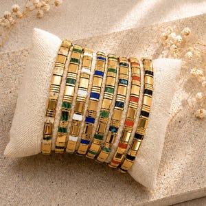 May include: A collection of eight gold-toned metal bracelets arranged side-by-side on a small rectangular pillow. Each bracelet features a series of rectangular links alternating between polished gold metal and colourful enamel or stone-like segments.
