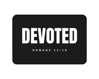 Puede incluir: Una alfombrilla de escritorio rectangular negra con la palabra DEVOTED en letras mayúsculas blancas grandes centradas. Debajo del texto principal, la referencia ROMANS 12:10 está impresa en una fuente sans-serif blanca más pequeña sobre el fondo negro.