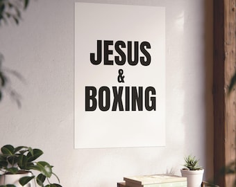 Puede incluir: Un póster de pared rectangular blanco que presenta el texto negro JESUS & BOXING en una fuente negrita sin remates. El póster se muestra verticalmente en una pared lisa, centrado sobre una superficie de madera con una planta pequeña.