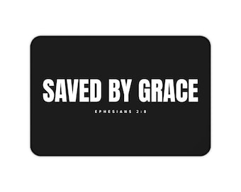 Puede incluir: Un tapete rectangular negro que presenta el texto blanco SAVED BY GRACE en una fuente sans-serif en negrita, con el texto blanco más pequeño EPHESIANS 2:8 colocado directamente debajo de la frase principal, todo centrado en el fondo oscuro.