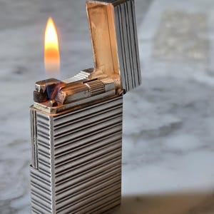 Könnte beinhalten: Ein rechteckiges Feuerzeug aus silberfarbenem Metall mit vertikaler Rillenstruktur. Das Feuerzeug ist geöffnet, der Deckel steht aufrecht, und eine kleine, hellorangefarbene Flamme brennt an der oberen Brennerbaugruppe.