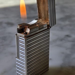 Könnte beinhalten: Ein rechteckiges Feuerzeug aus silberfarbenem Metall mit einer vertikal gerippten Textur auf dem Gehäuse und dem Deckel. Der Deckel ist geöffnet und gibt den Blick auf den Zündmechanismus frei, während eine kleine, helle orangefarbene Flamme aufsteigt.