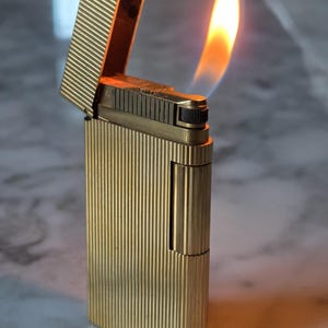 Könnte beinhalten: Ein rechteckiges Feuerzeug aus goldfarbenem Metall mit vertikaler Nadelstreifen-Textur. Der Klappdeckel ist geöffnet und zeigt eine brennende Flamme. Der Zündmechanismus verfügt über ein kleines, schwarzes, geriffeltes Rädchen.