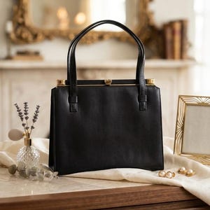 May include: A black handbag with two handles and gold-colored accents. The rectangular purse is sitting on a wooden surface. A small perfume bottle, a gold picture frame, and a pair of pearl earrings are also on the surface.