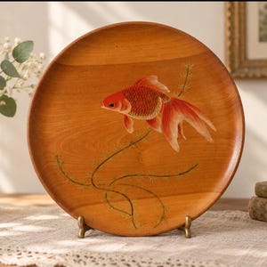 May include: A round wooden plate featuring a painted orange and red goldfish swimming among green aquatic plants. The plate has a smooth, polished finish with visible wood grain patterns and is displayed upright on a small metal stand.