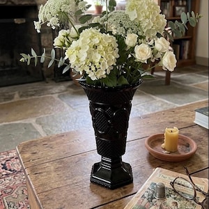 May include: A black glass vase with a textured diamond pattern and a flared rim, containing a bouquet of white hydrangeas, small white roses, green eucalyptus sprigs, and white lace flowers, standing on a wooden surface.