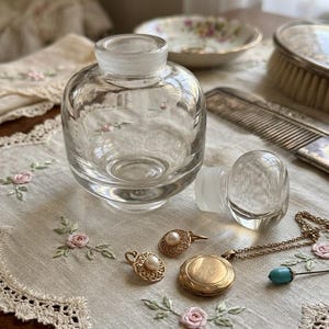 May include: A clear glass perfume bottle with a detached stopper sits on a floral embroidered cloth. Gold-toned jewelry, including earrings, a locket, and a necklace, are arranged nearby. A comb and brush are in the background.