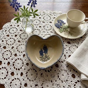 May include: A heart-shaped ceramic bowl with a speckled beige finish and a blue lupine flower illustration at the center. The bowl features a dark blue rim and is positioned on a white crocheted doily alongside a cup and saucer with flowers.