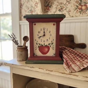 May include: A rectangular wooden mantel clock with a dark green base and top, red side pillars, and a cream-colored face. The clock face features black Arabic numerals, black hands, and a hand-painted red apple illustration with green leaves.