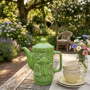 May include: A light green ceramic teapot featuring a raised relief pattern of a mushroom, flowers, and a butterfly over a woven basket texture. The teapot has a matching lid, a curved spout, and a handle, accompanied by two stacked cream-colored cups.