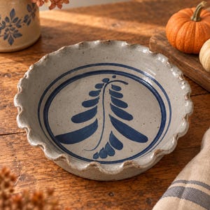 May include: A round ceramic pie dish with a speckled light gray finish and a ruffled edge. The interior features a hand-painted dark blue fern-like botanical design enclosed within a circular border, showcasing a classic stoneware aesthetic.