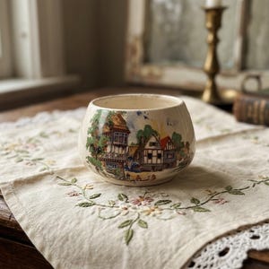 May include: A cream-colored ceramic bowl featuring a colorful transferware illustration of a village scene with timber-framed houses, green trees, and a blue sky. The bowl has a rounded shape with a wide opening and a smooth, glossy finish.