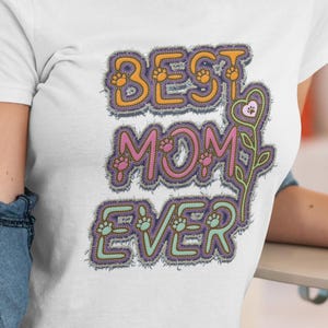 以下が含まれることがあります： カラフルなステッチデザインで「BEST MOM EVER」の文字が入った白いTシャツ。テキストには足跡と花のアクセントが含まれています。このシャツは半袖で、柔らかい素材で作られているようです。