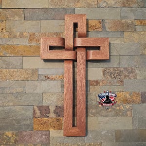 Op de afbeelding: Een houten kruis met een geweven ontwerp, gepresenteerd tegen een veelkleurige stenen muur. Het kruis is gemaakt van roodbruin hout en heeft een uniek in elkaar grijpend patroon. Een sticker "Veteran Owned Business" is zichtbaar.
