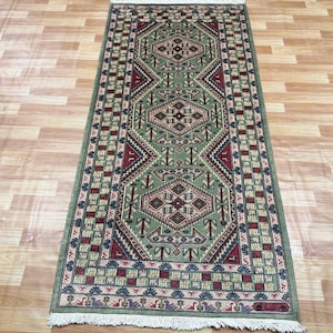 May include: A rectangular runner rug featuring a sage green field with three central diamond-shaped medallions in red, cream, and dark blue. The border displays a repeating geometric pattern with fringe on both ends of the woven textile.