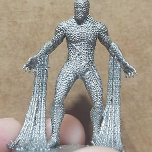 May include: A silver figurine of a humanoid figure with outstretched arms and flowing fabric extensions. The figure has a textured, scale-like appearance. The figurine is standing on a small, round base.